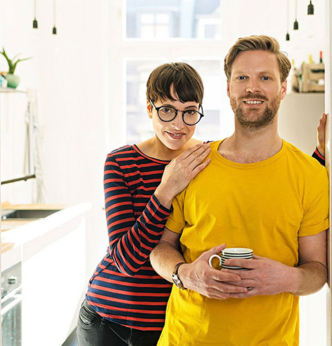 A young man and a woman standing in a brightly lit kitchen. The man is holding a cup of coffee. 