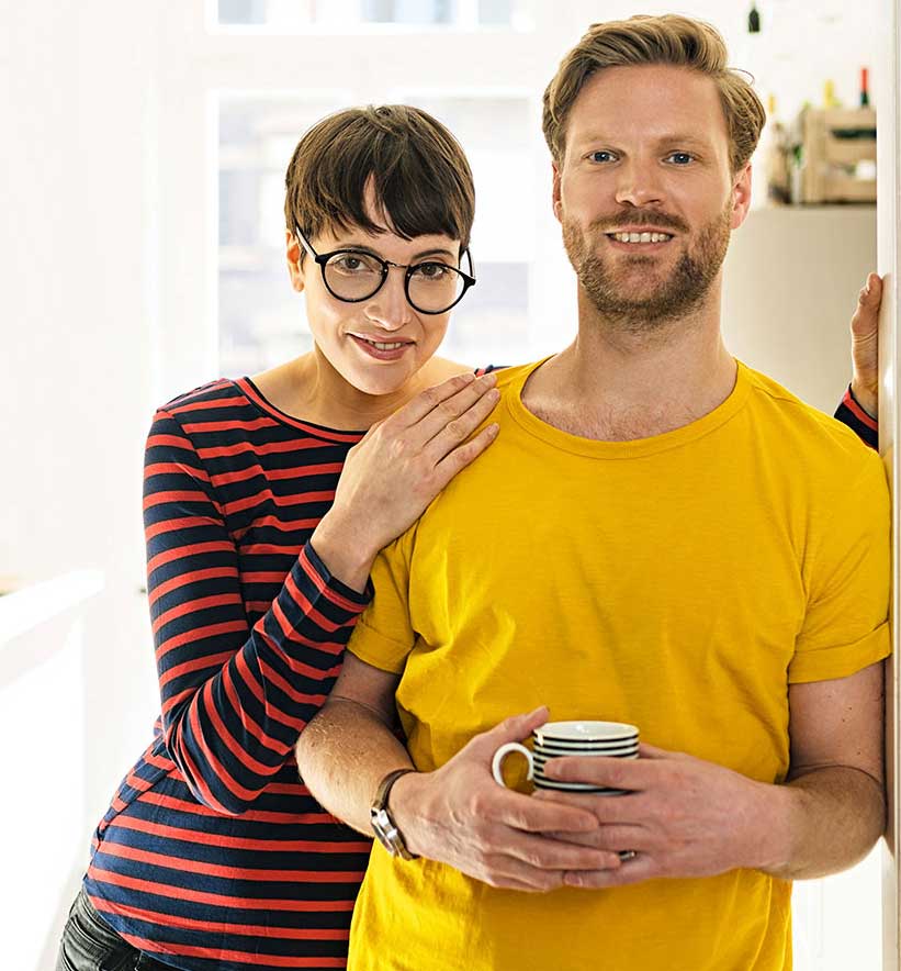 A young man and a woman standing in a brightly lit kitchen. The man is holding a cup of coffee. 