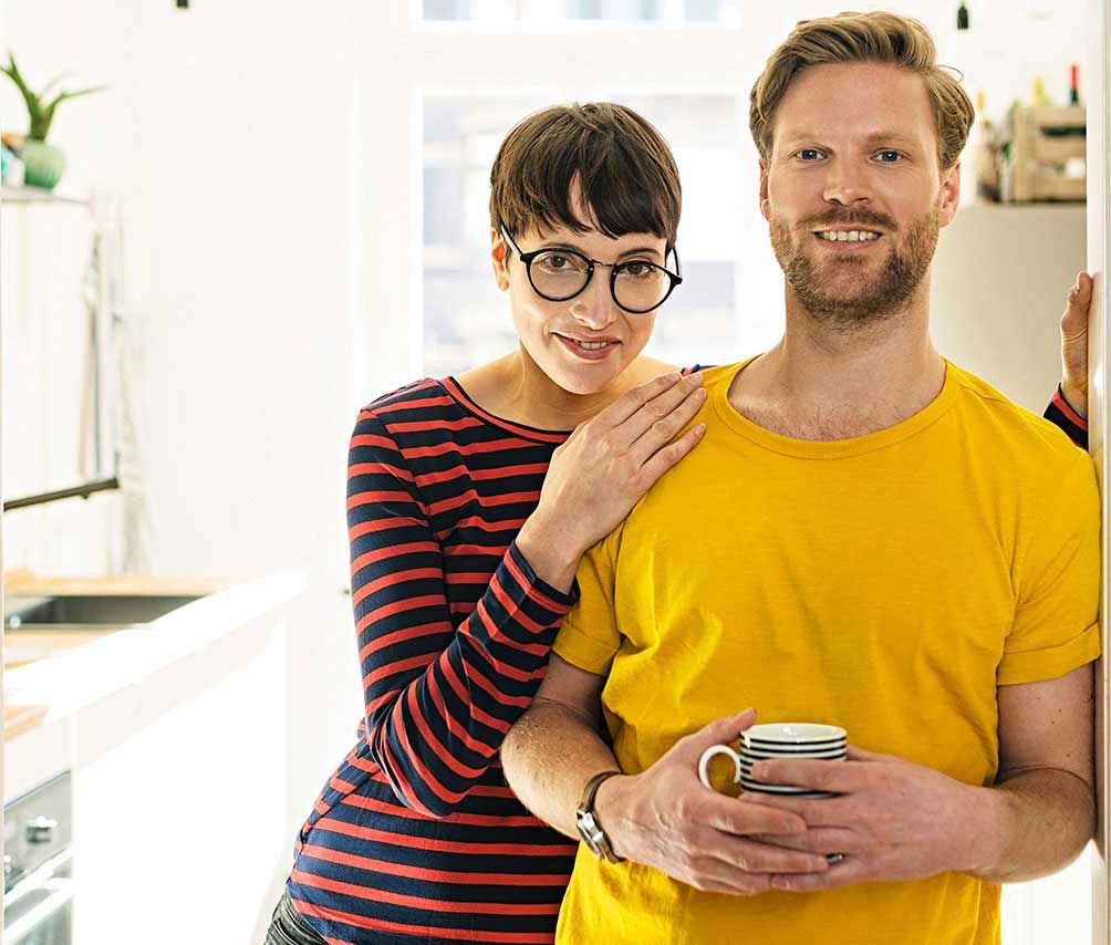 A young man and a woman standing in a brightly lit kitchen. The man is holding a cup of coffee. 