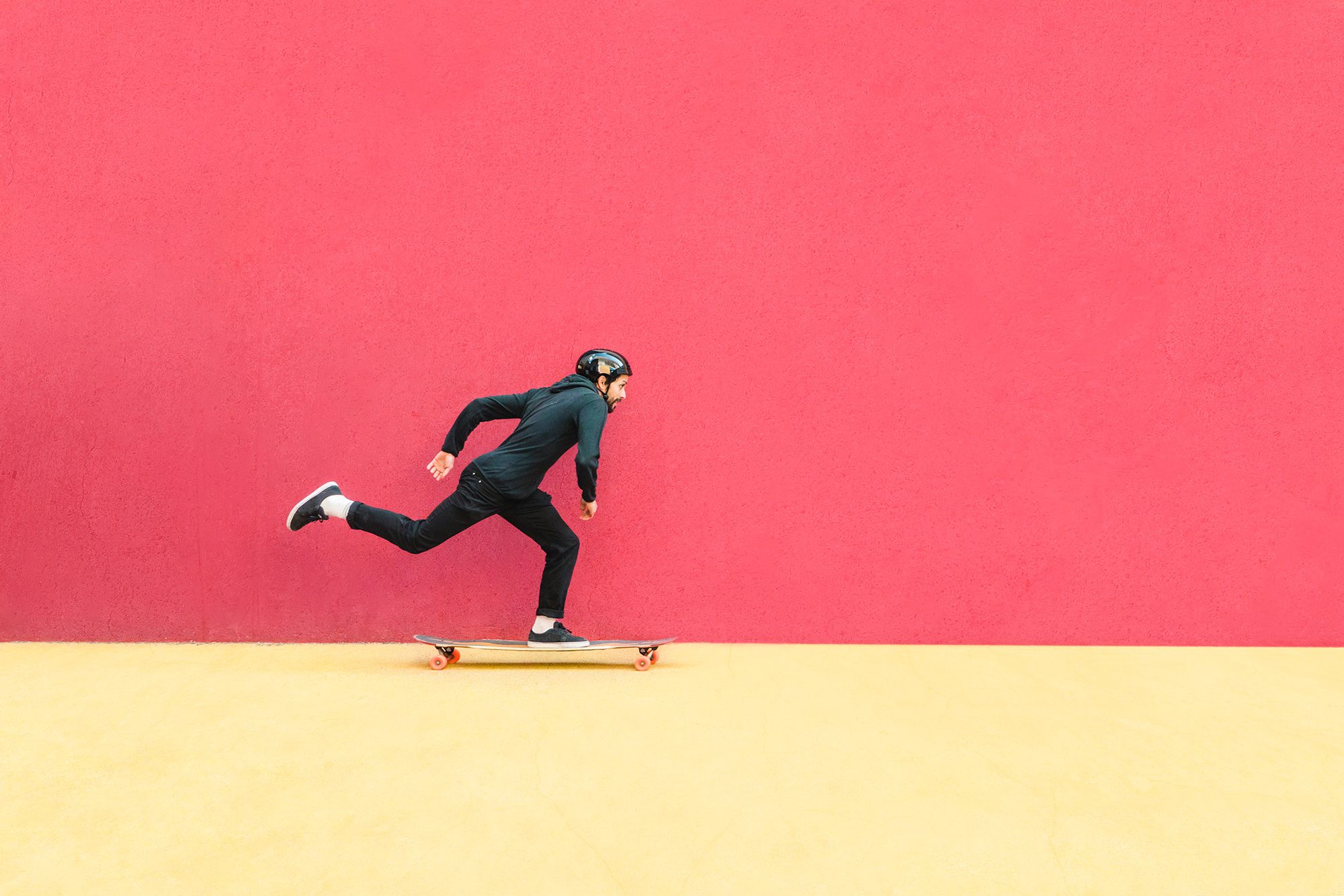 A young man with a beard skateboarding in front of a red wall